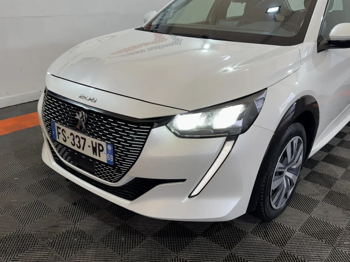 Vue 3/4 avant droit d'une Peugeot 208 électrique blanche avec phares LED allumés et calandre noire distinctive.