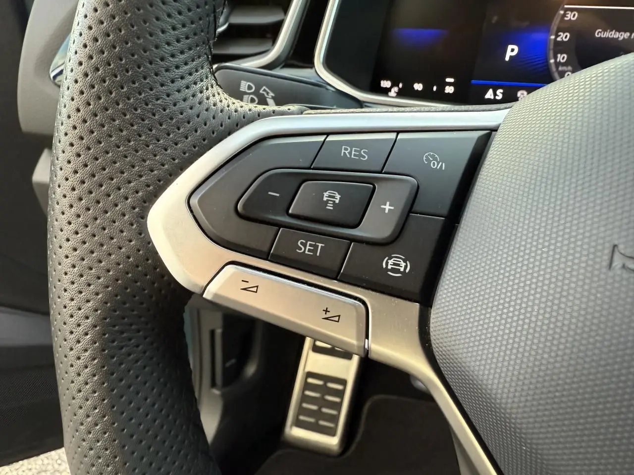 Gros plan sur les commandes multifonctions à gauche du volant perforé d’un Volkswagen T-Roc 2025, avec pédale en acier visible.