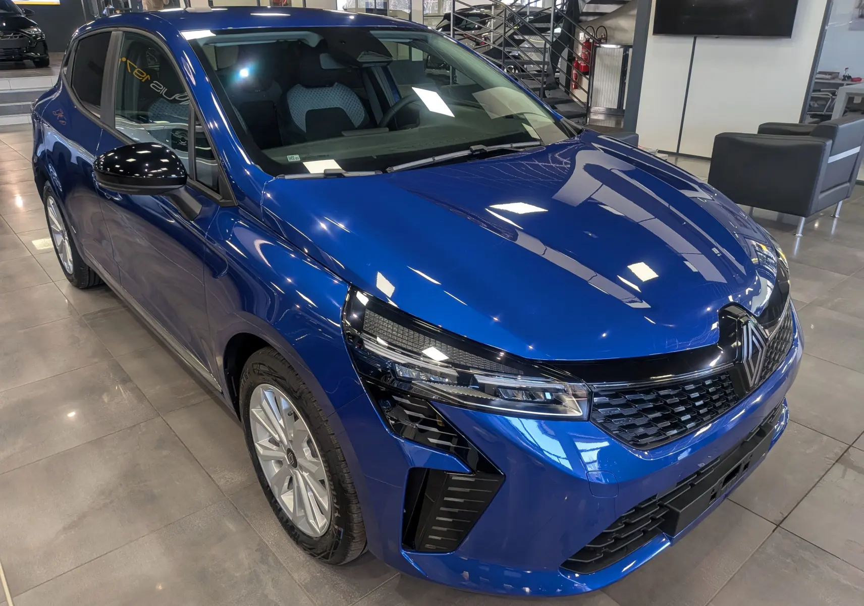 Vue 3/4 avant gauche de la Renault Clio V bleu Iron avec calandre noire et rétroviseurs noirs brillants en showroom.