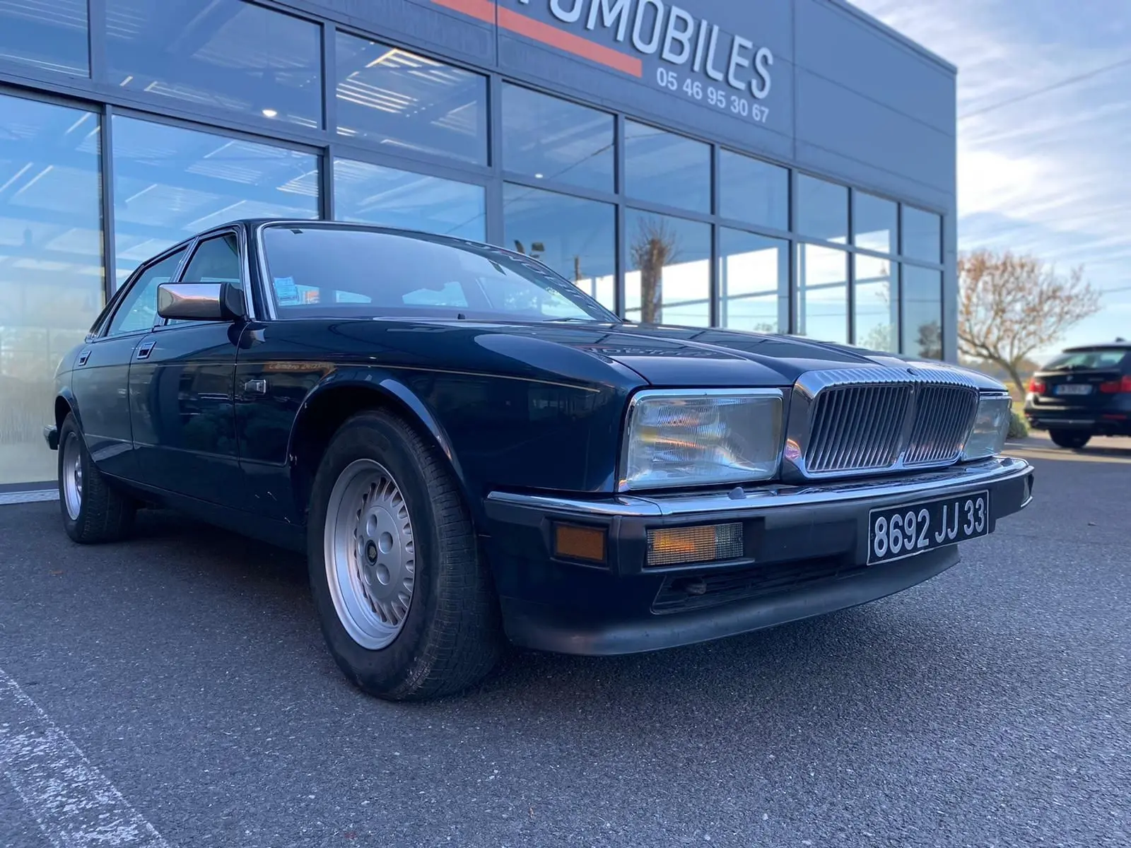Jaguar XJ6 3.6 Sovereign bleu vue 3/4 avant avec calandre chromée et phares rectangulaires, stationnée devant un showroom.