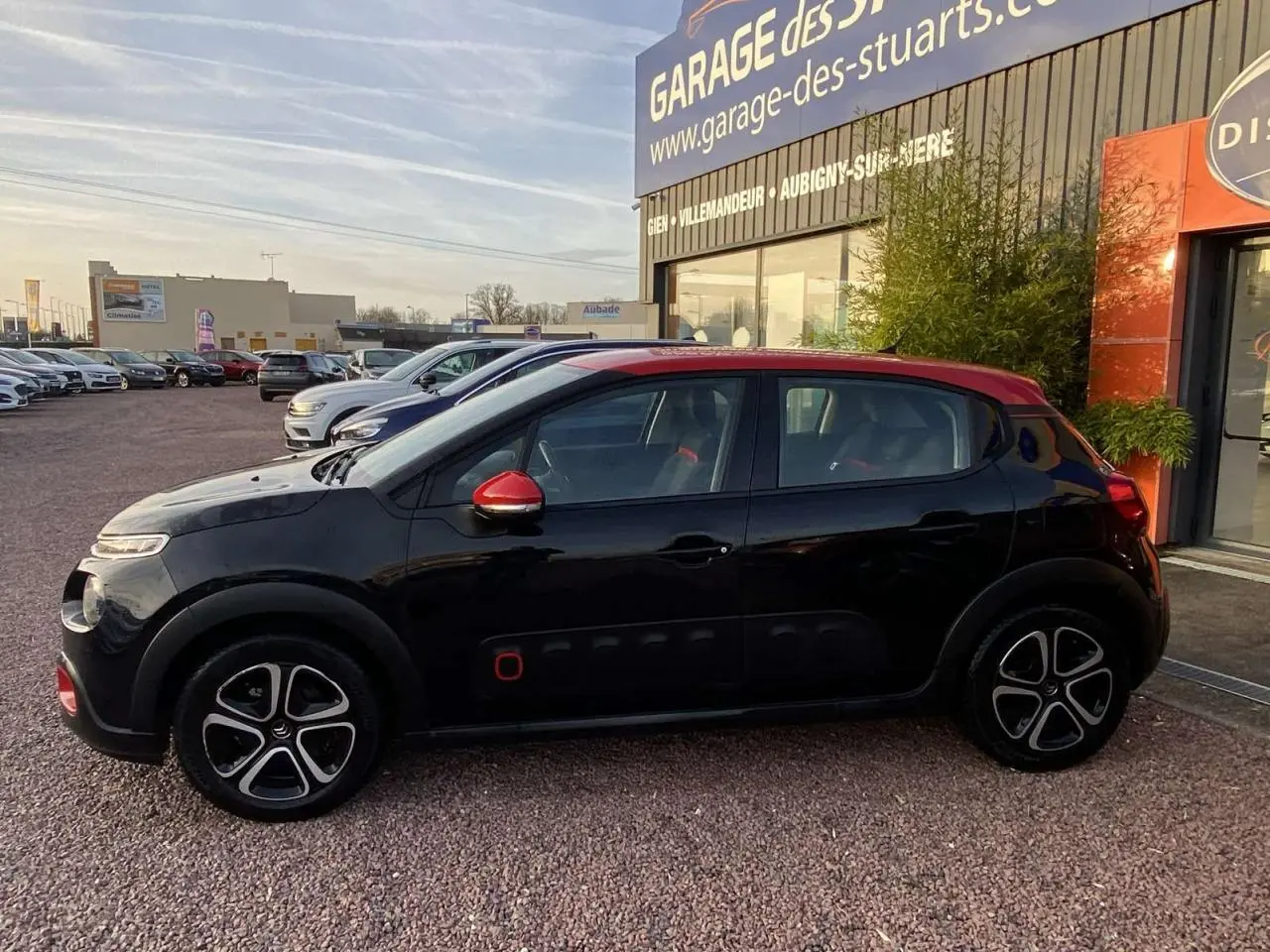 Profil côté gauche d'une Citroën C3 noire avec toit et rétroviseurs rouges, garée devant un garage.