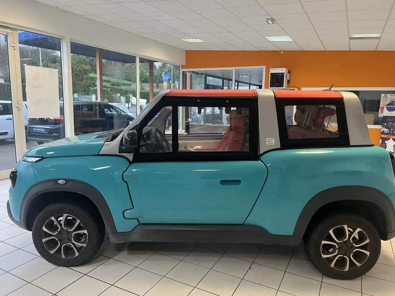 Vue de profil côté gauche d'une Citroën E-MEHARI cabriolet bleu clair avec toit en toile rouge en intérieur showroom.