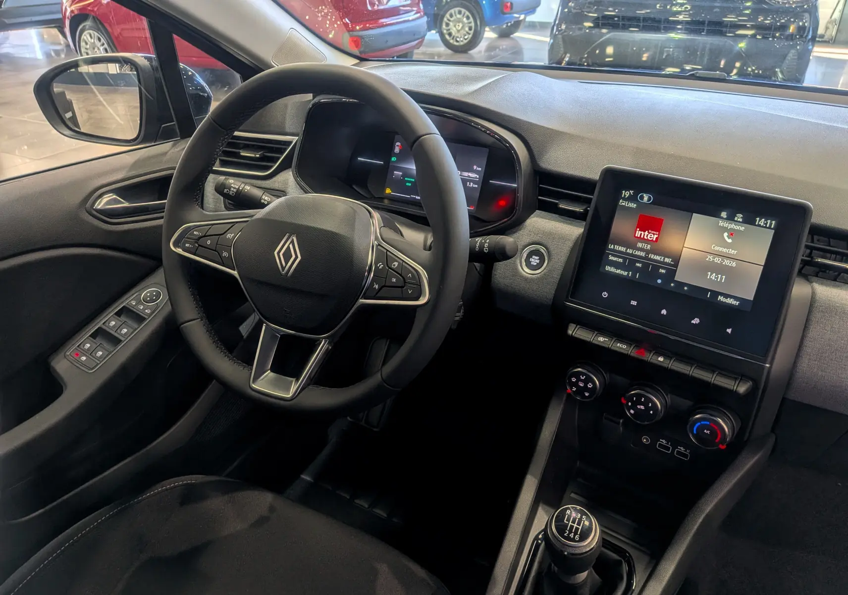 Vue intérieure de face du poste de conduite de la Renault Clio V avec volant, écran tactile et levier de vitesses manuel.