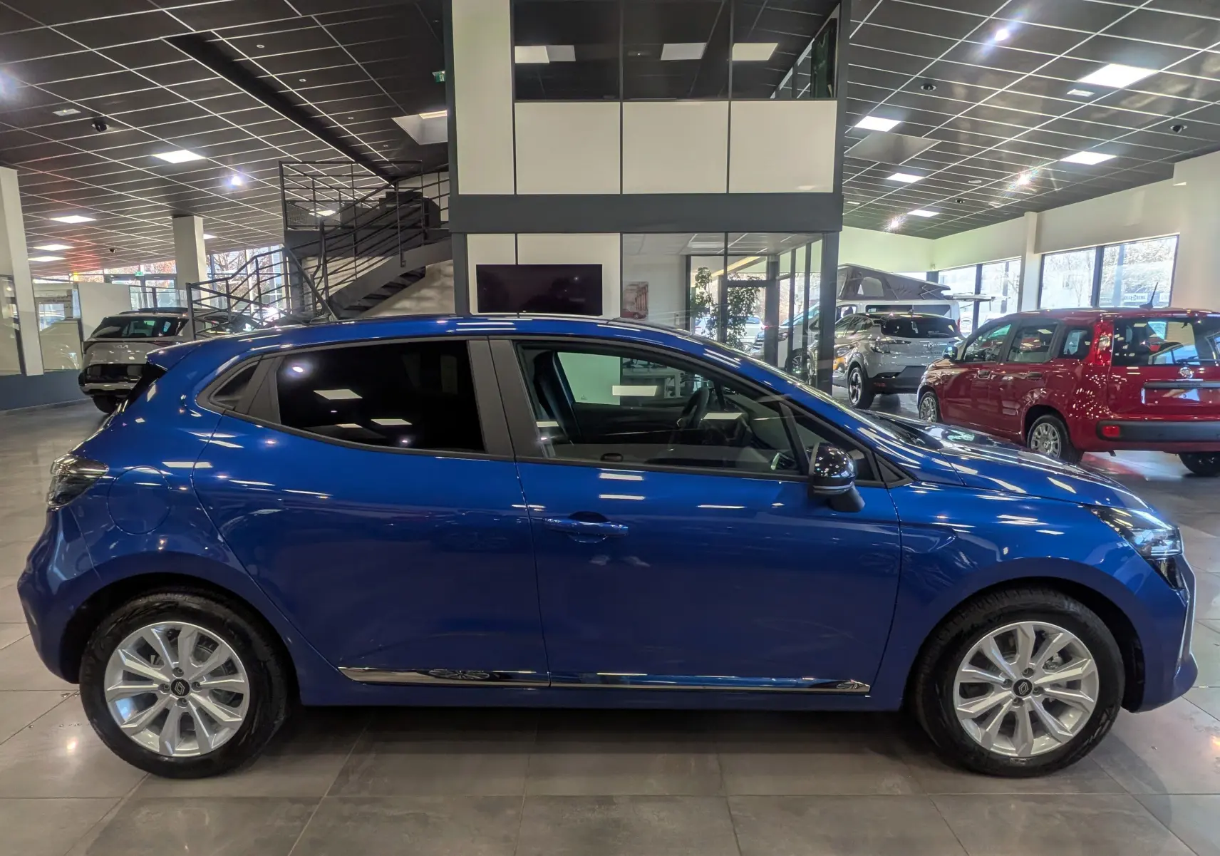 Vue de profil côté gauche d'une Renault Clio V Bleu Iron 2025 en intérieur, avec jantes alliage et vitres teintées.