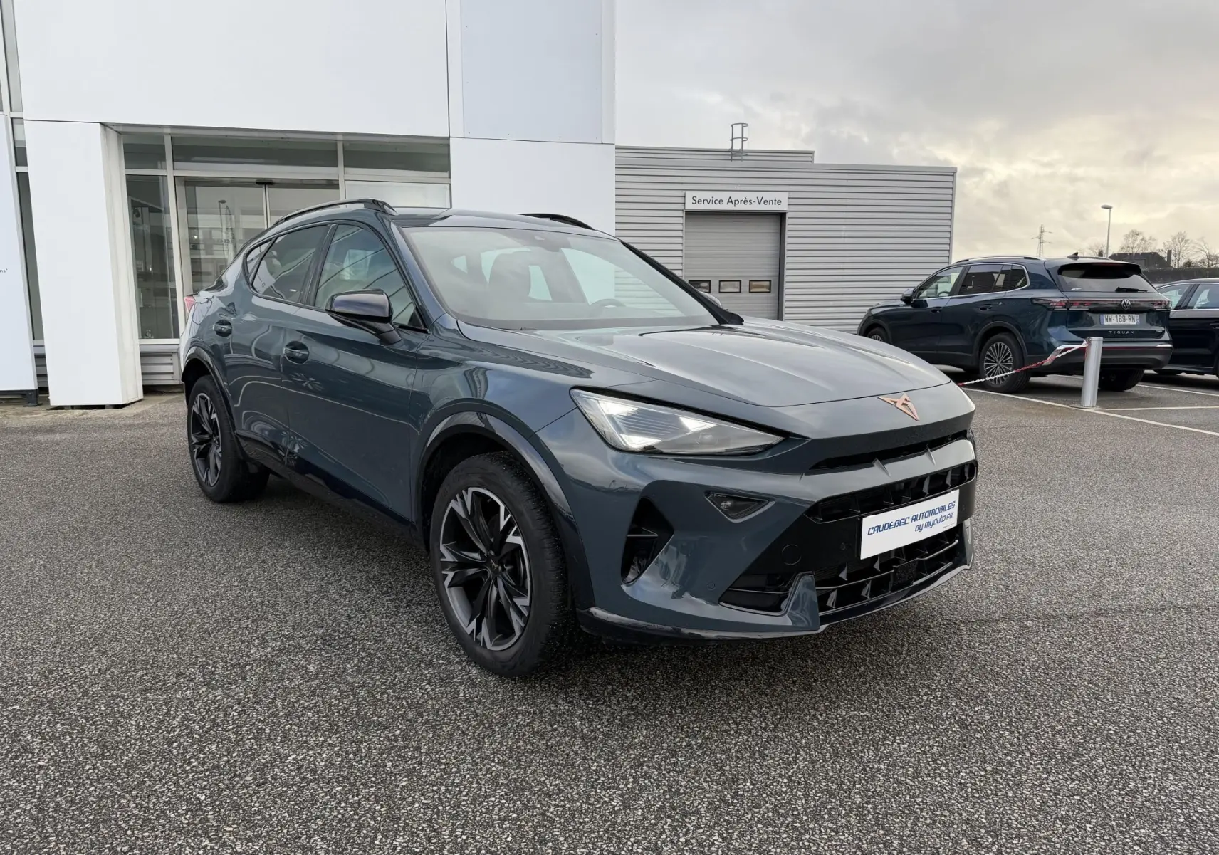 Vue 3/4 avant droit d'un CUPRA Formentor bleu métallisé avec jantes noires, garé devant un bâtiment commercial.