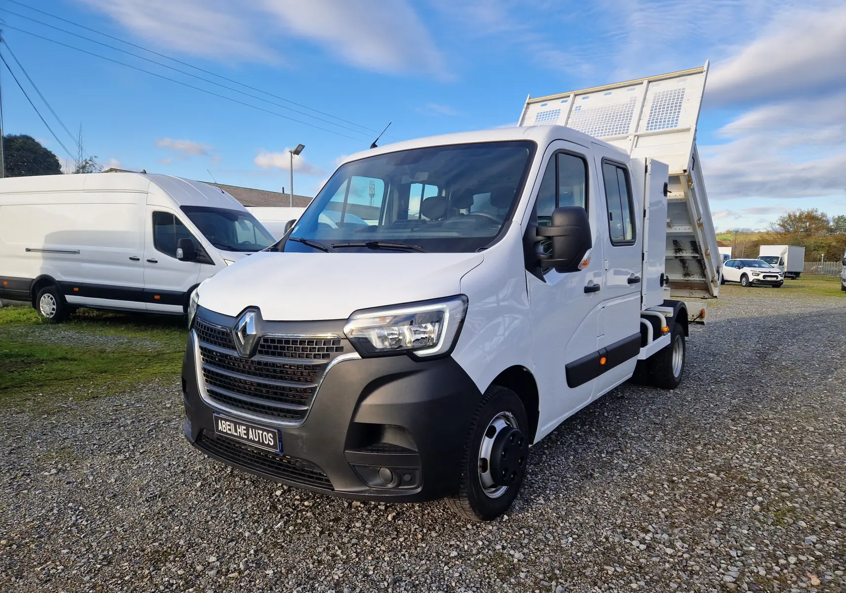 Renault Master blanc en 3/4 avant droit avec benne relevée et coffre latéral sur terrain gravillonné.