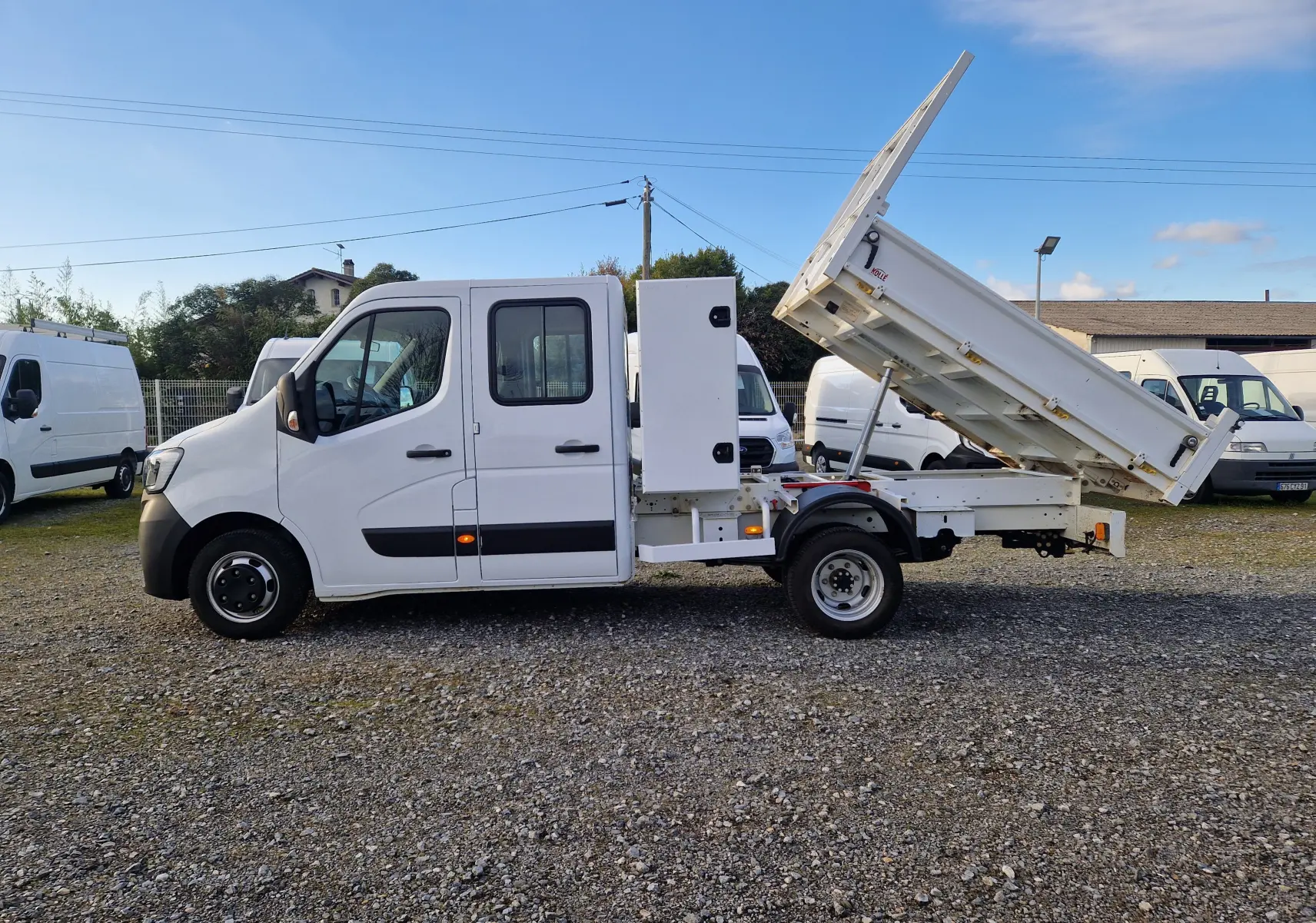 Profil droit d'un Renault Master Châssis double cabine blanc avec benne relevée et coffre latéral visible sur un terrain extérieur.