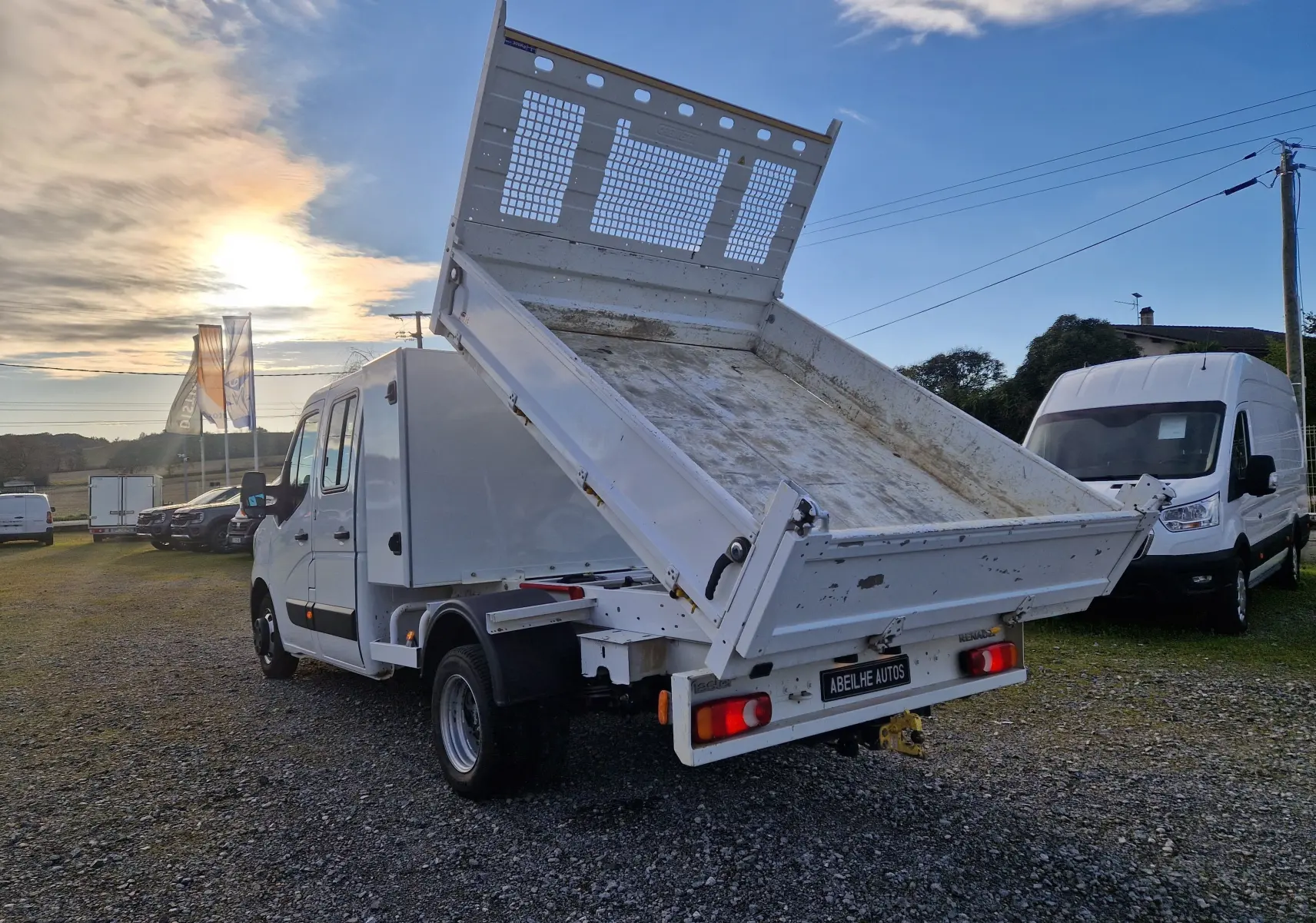 Renault Master blanc vu de l'arrière droit avec benne relevée et cabine double, stationné en extérieur au coucher du soleil.