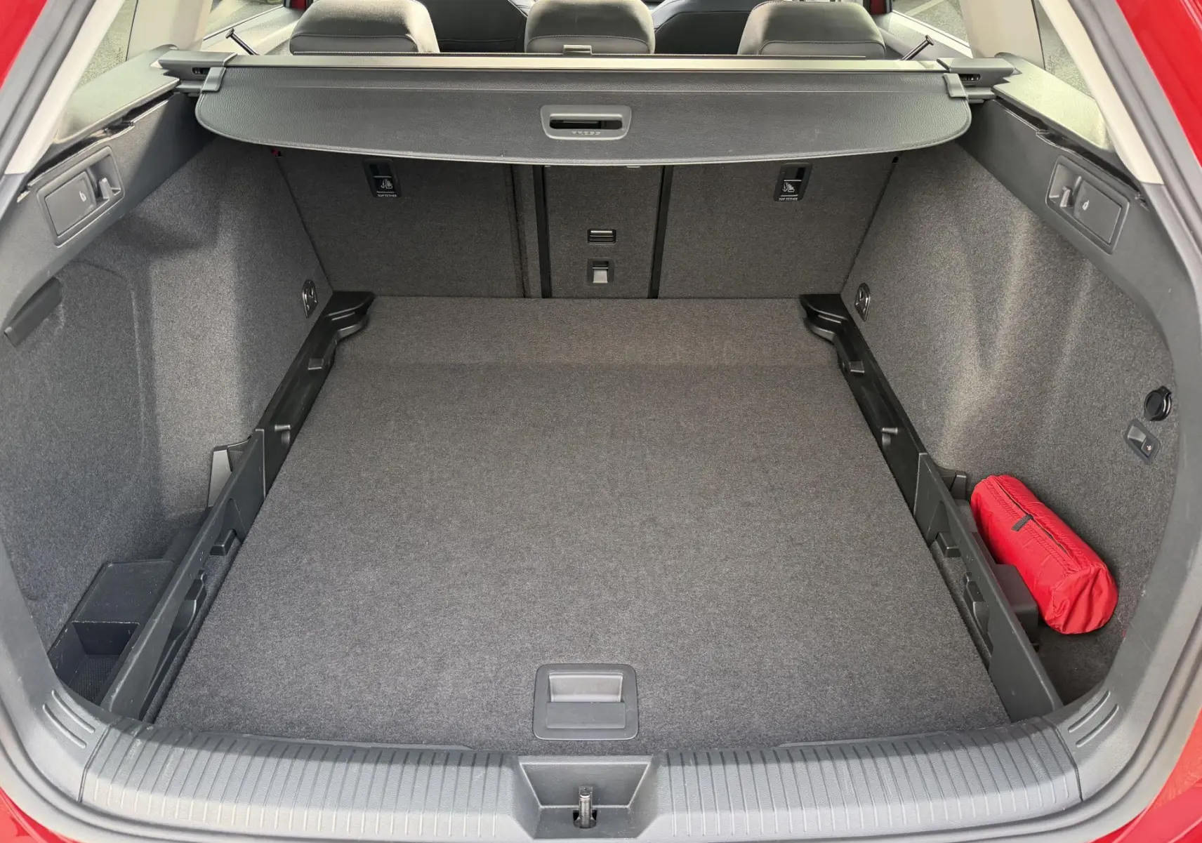 Coffre ouvert vu de l'arrière d'une Volkswagen Golf SW rouge, avec tapis gris et trousse de secours rouge à droite.