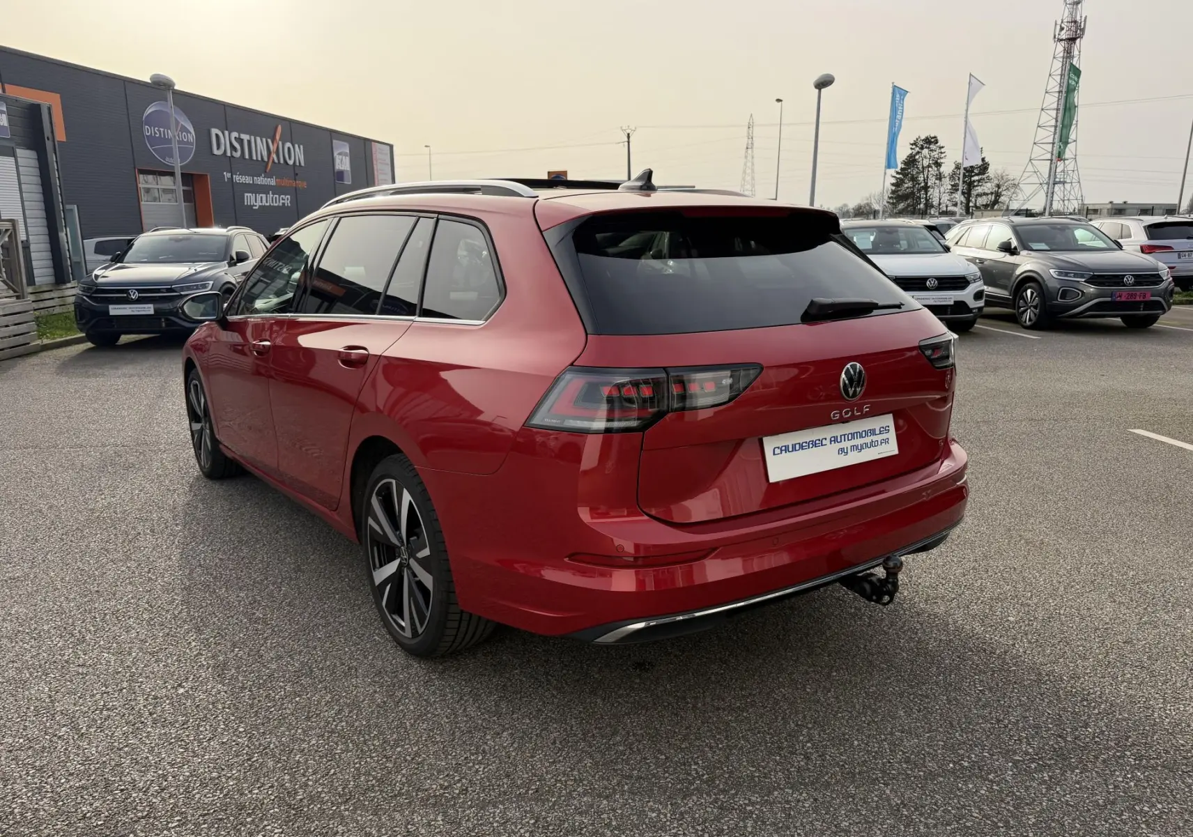 Vue 3/4 arrière droite d'une Volkswagen Golf SW rouge métallisée avec attelage rabattable sur parking extérieur.
