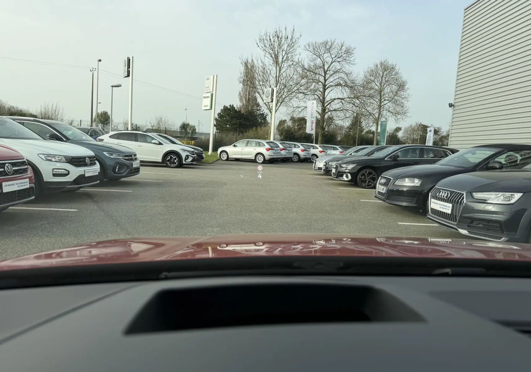 Vue depuis l'habitacle d'une Volkswagen Golf SW rouge, avec le capot visible au premier plan, dans un parking extérieur.
