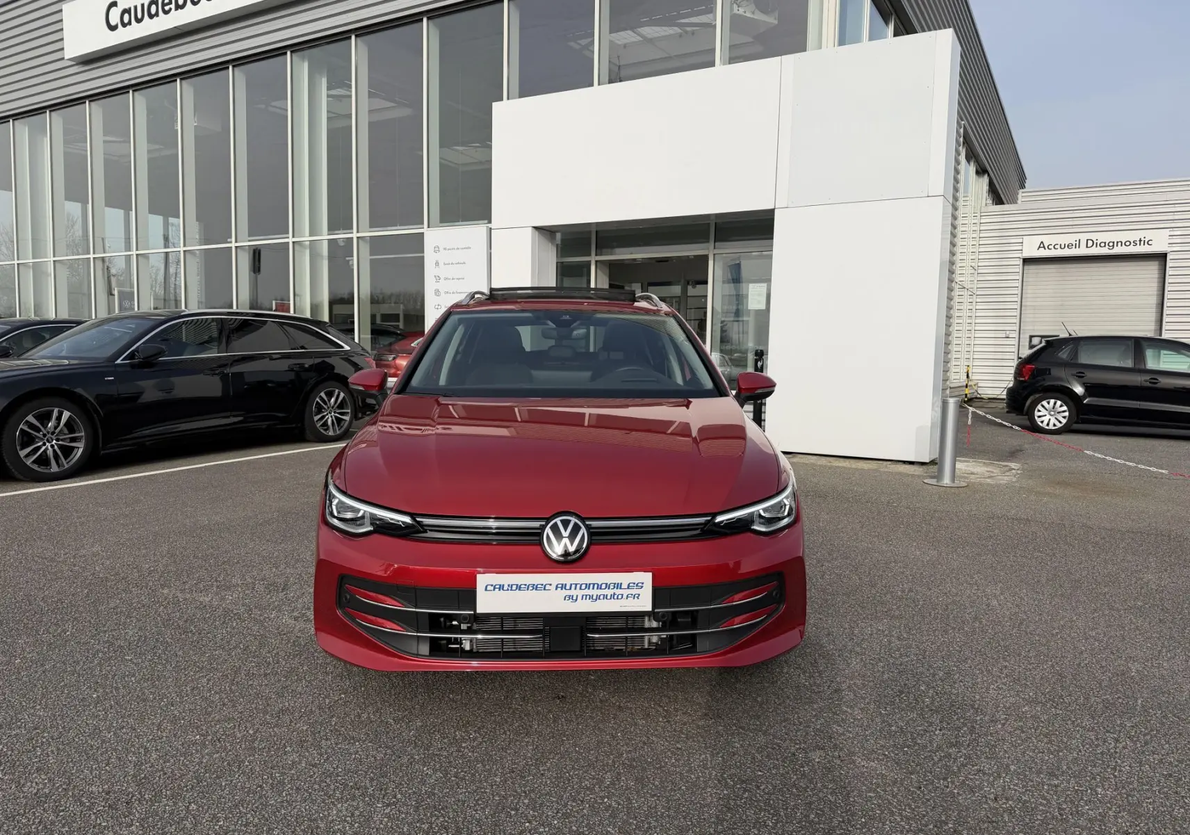 Vue frontale d'une Volkswagen Golf SW rouge métallisée garée devant un bâtiment commercial.