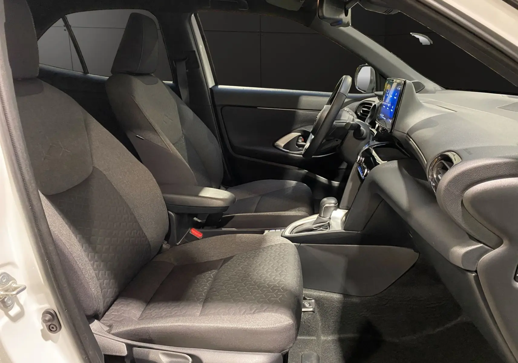 Vue intérieure côté conducteur de la Toyota Yaris Cross blanche, sièges noirs en tissu et tableau de bord avec écran tactile.