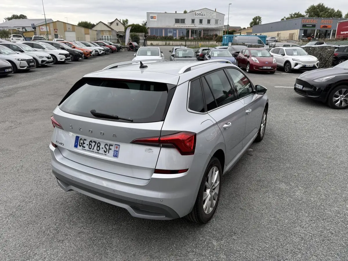 Vue 3/4 arrière droite d'un Skoda Kamiq gris métallisé avec barres de toit et feux arrière LED dans un parking.