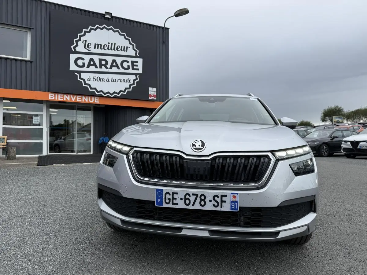 Vue de face d'un Skoda Kamiq gris métallisé avec calandre noire et phares LED allumés devant un garage.