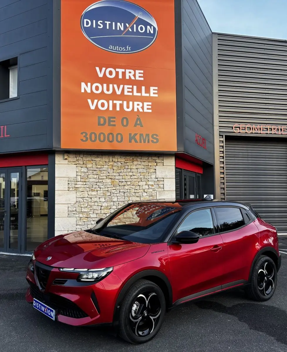 Vue 3/4 avant droite d'une Alfa Romeo Junior rouge avec toit noir, jantes noires et calandre distinctive devant un bâtiment commercial.