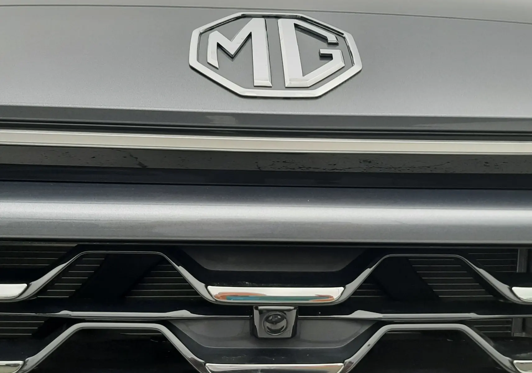 Gros plan sur la calandre avant grise Camden du MG ZS 1.5 HYBRID+ 197CH avec logo MG et caméra frontale intégrée.