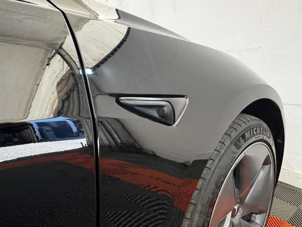 Gros plan sur l'aile avant droite noire d'une Tesla Model 3 2023 avec caméra latérale intégrée et jante grise.