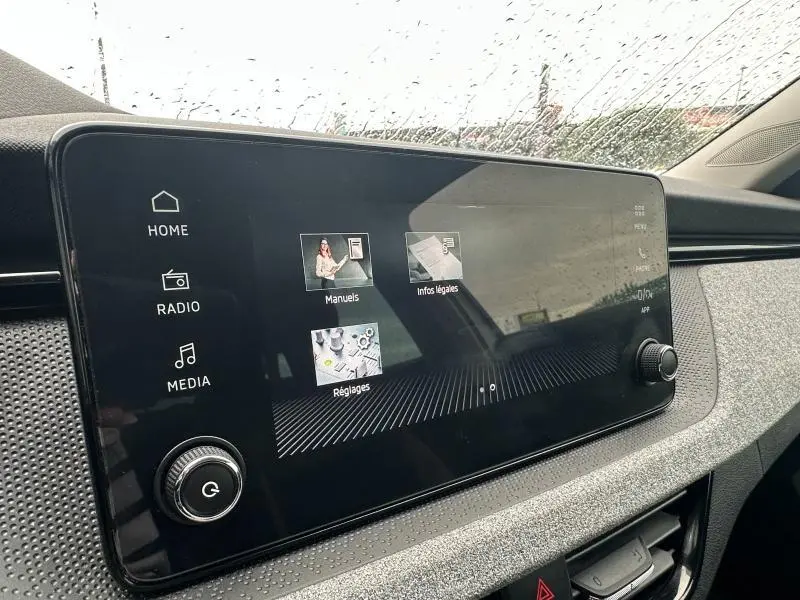 Écran tactile central du tableau de bord du Skoda Scala blanc, avec commandes multimédia visibles sous la pluie.