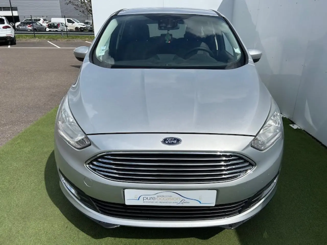 Vue avant d'une Ford C-MAX II gris clair métal avec calandre chromée et phares halogènes, stationnée sur sol vert.