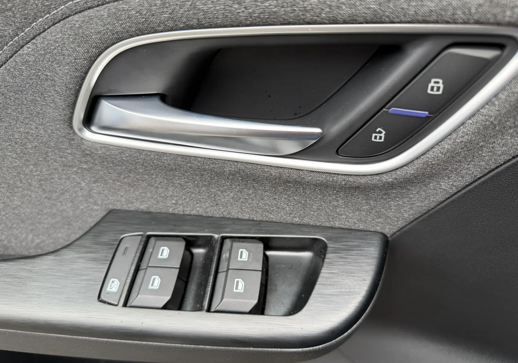 Poignée de porte intérieure et commandes des vitres en tissu gris sur MG ZS 1.5L Hybrid+ 2026, détails en plastique noir et argenté.