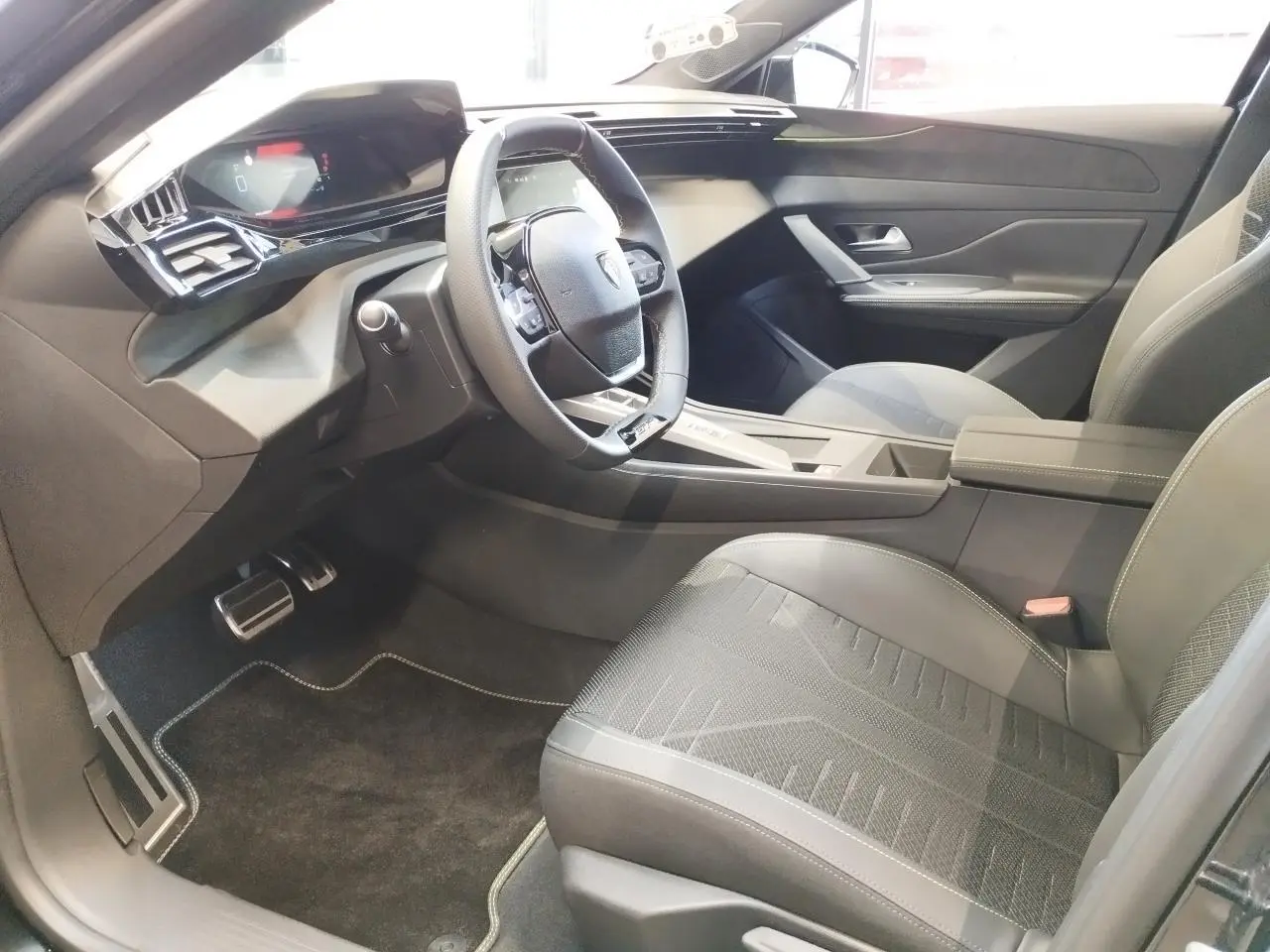 Vue intérieure côté conducteur de la Peugeot 308 1.2i Hybrid 2026, avec sièges noirs et volant compact GT cuir.
