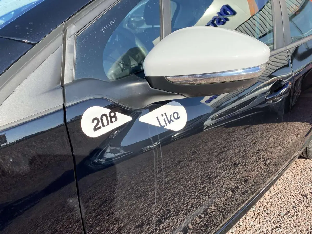Vue côté gauche rapprochée d'une Peugeot 208 noire avec coques de rétroviseurs blanches et stickers "208 Like" sur la porte.