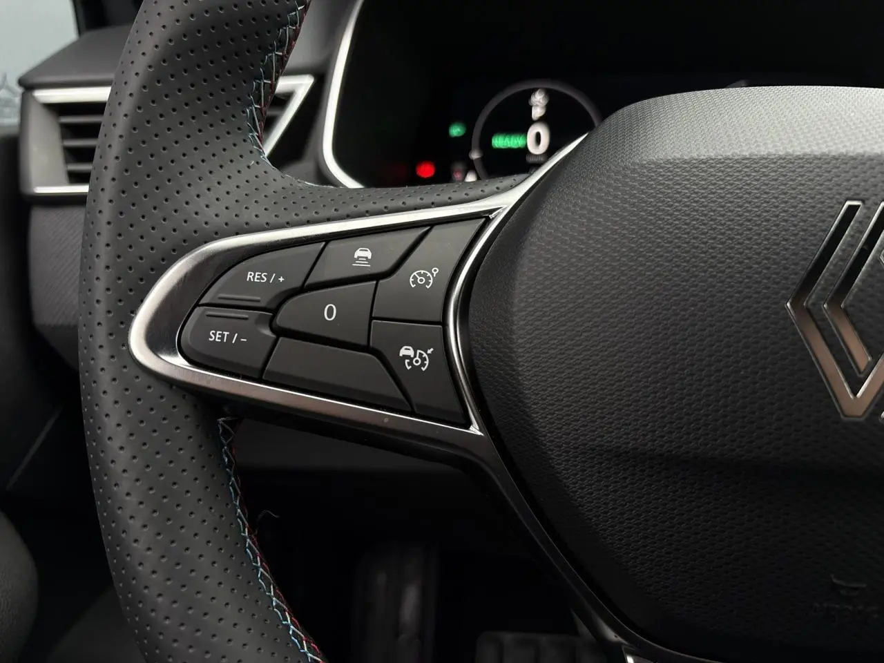 Gros plan sur les commandes du volant côté gauche d'une Renault Clio gris Rafale, avec surpiqûres bleues visibles.