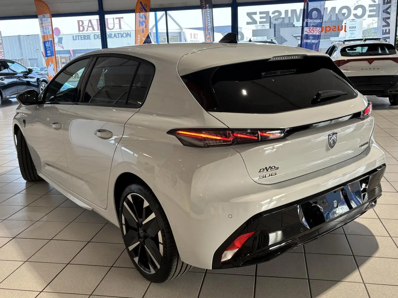 Vue 3/4 arrière droite d'une Peugeot 308 blanche hybride avec jantes bi-tons et bandeau noir brillant arrière en showroom.