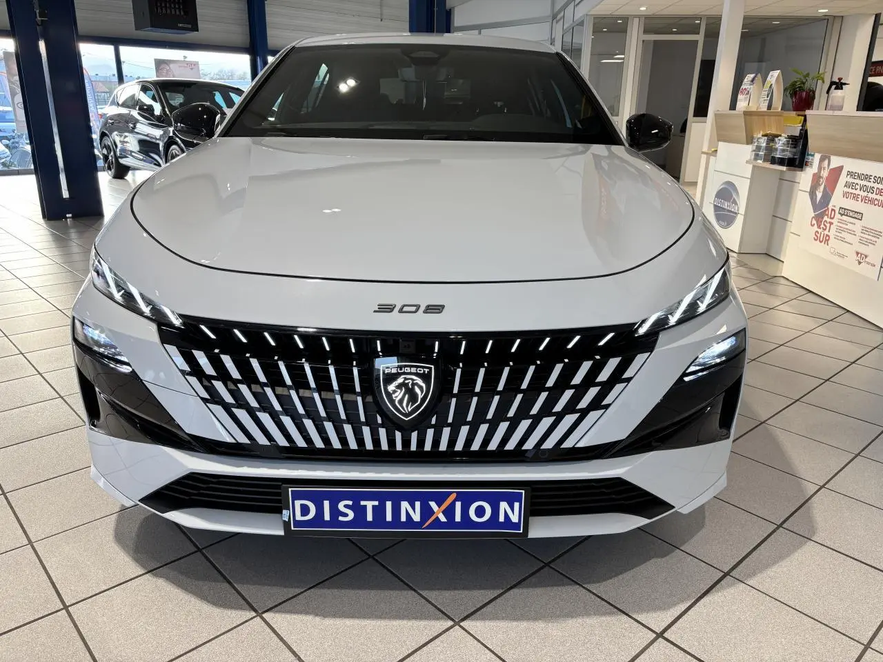 Vue de face d'une Peugeot 308 blanche avec calandre noire à motifs verticaux et logo lion lumineux, en showroom.