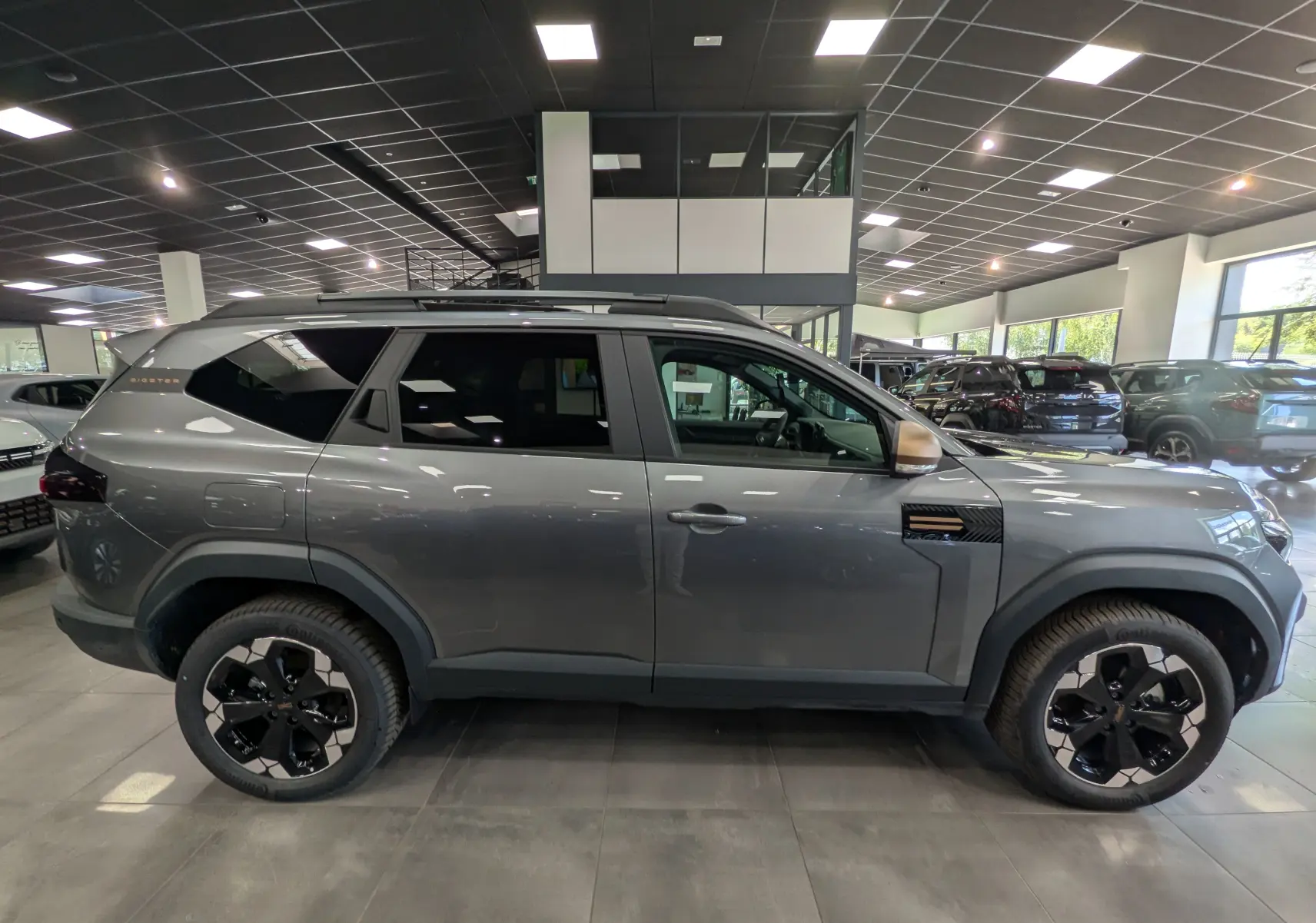 Vue de profil côté gauche du Dacia Bigster Hybrid-G 150 4x4 gris schiste avec jantes biton noires en showroom.
