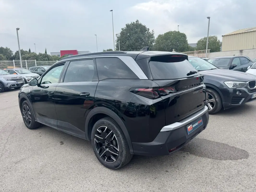 Vue 3/4 arrière droite d’un Peugeot 5008 noir avec jantes alliage noires et barres de toit visibles en extérieur.