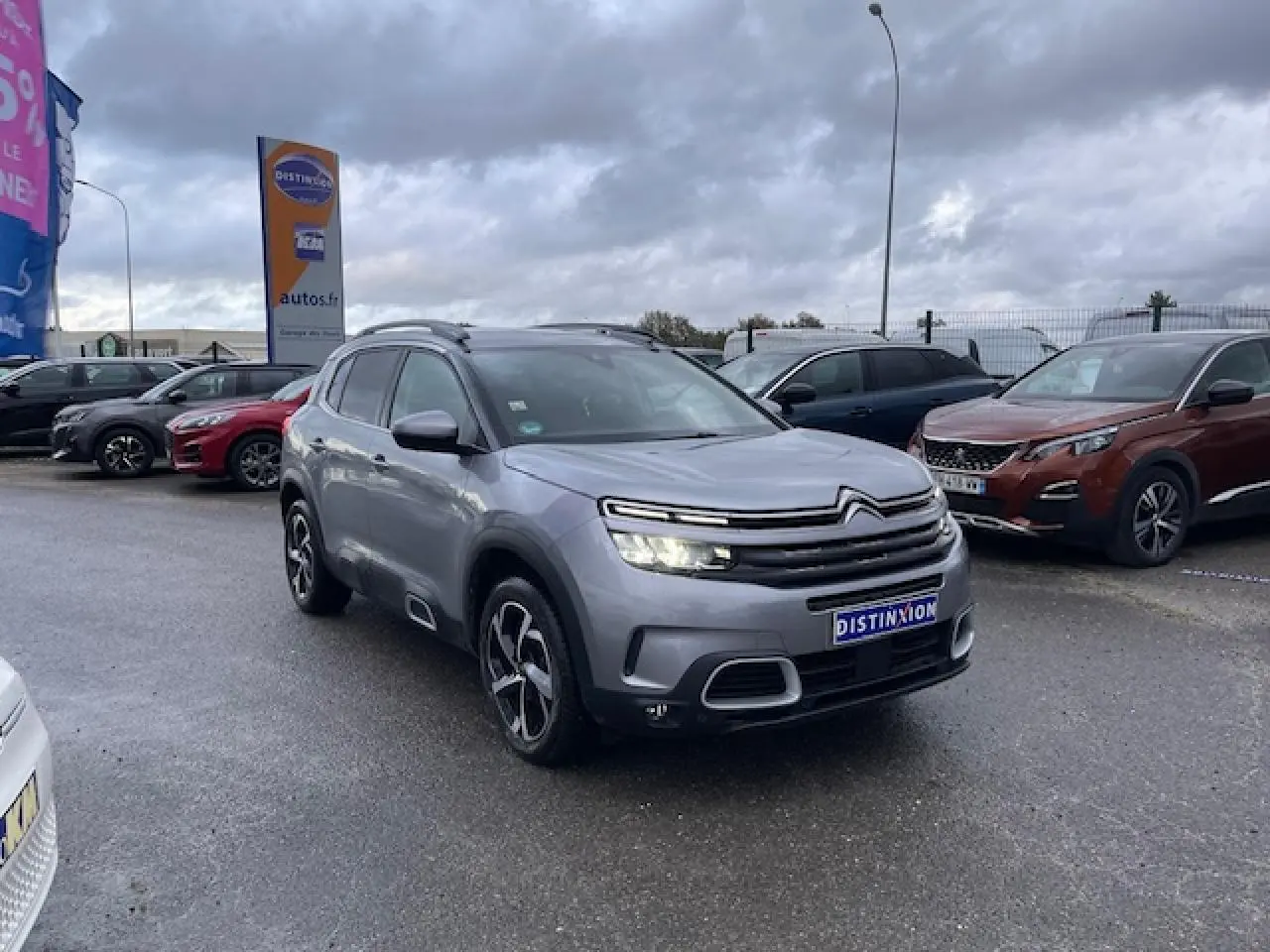 Citroën C5 Aircross gris Artense vue 3/4 avant avec feux allumés sur parking sous ciel nuageux.