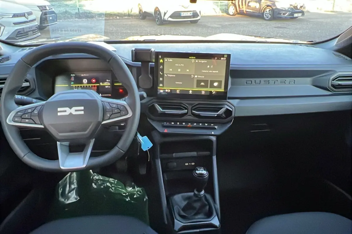 Vue intérieure du tableau de bord du Dacia Duster 2025, avec volant multifonction, écran tactile et levier de vitesse manuel.
