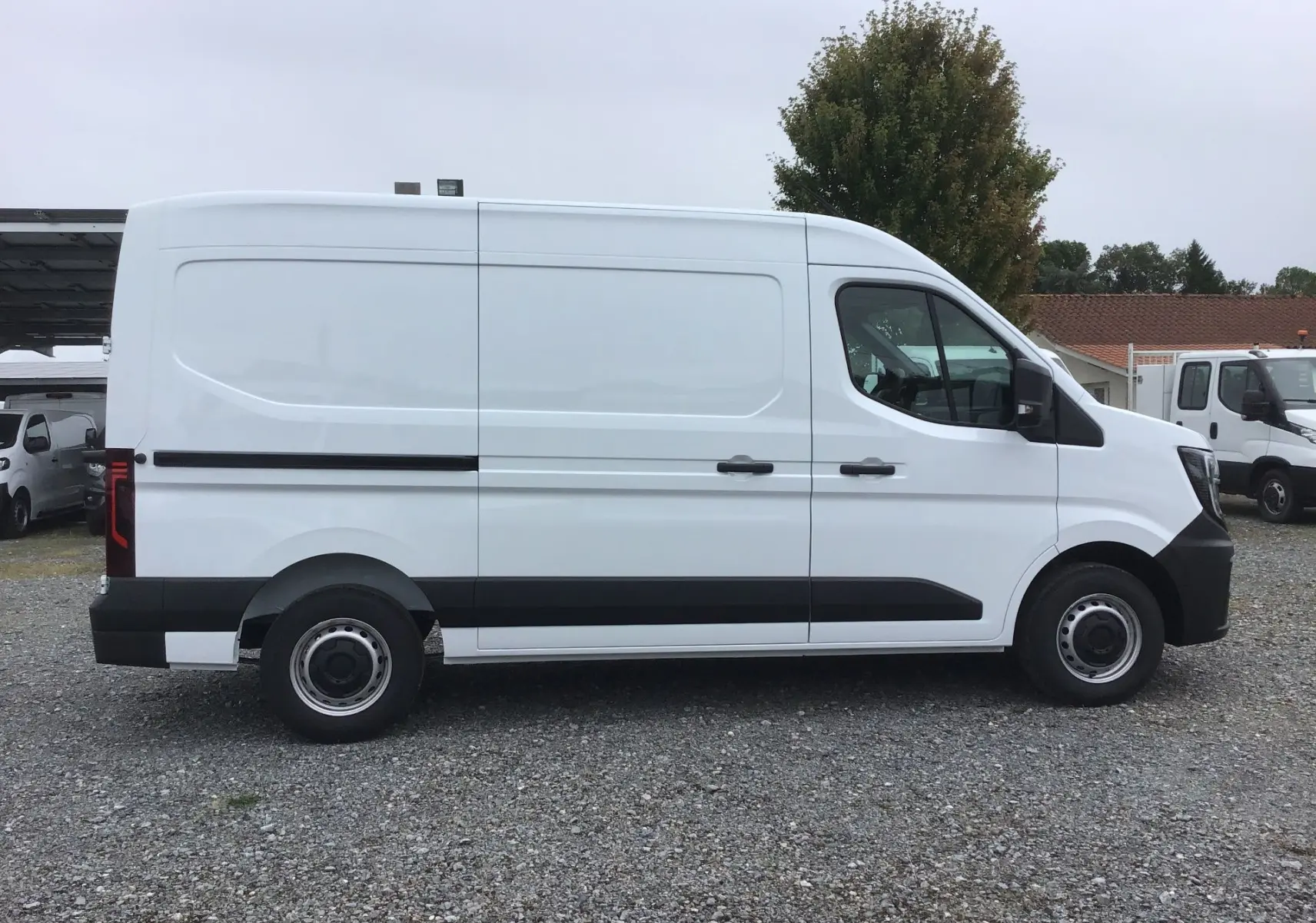 Vue de profil côté droit d’un Renault Master blanc L2H2 2025 avec bande noire et jantes acier sur un sol gravillonné.