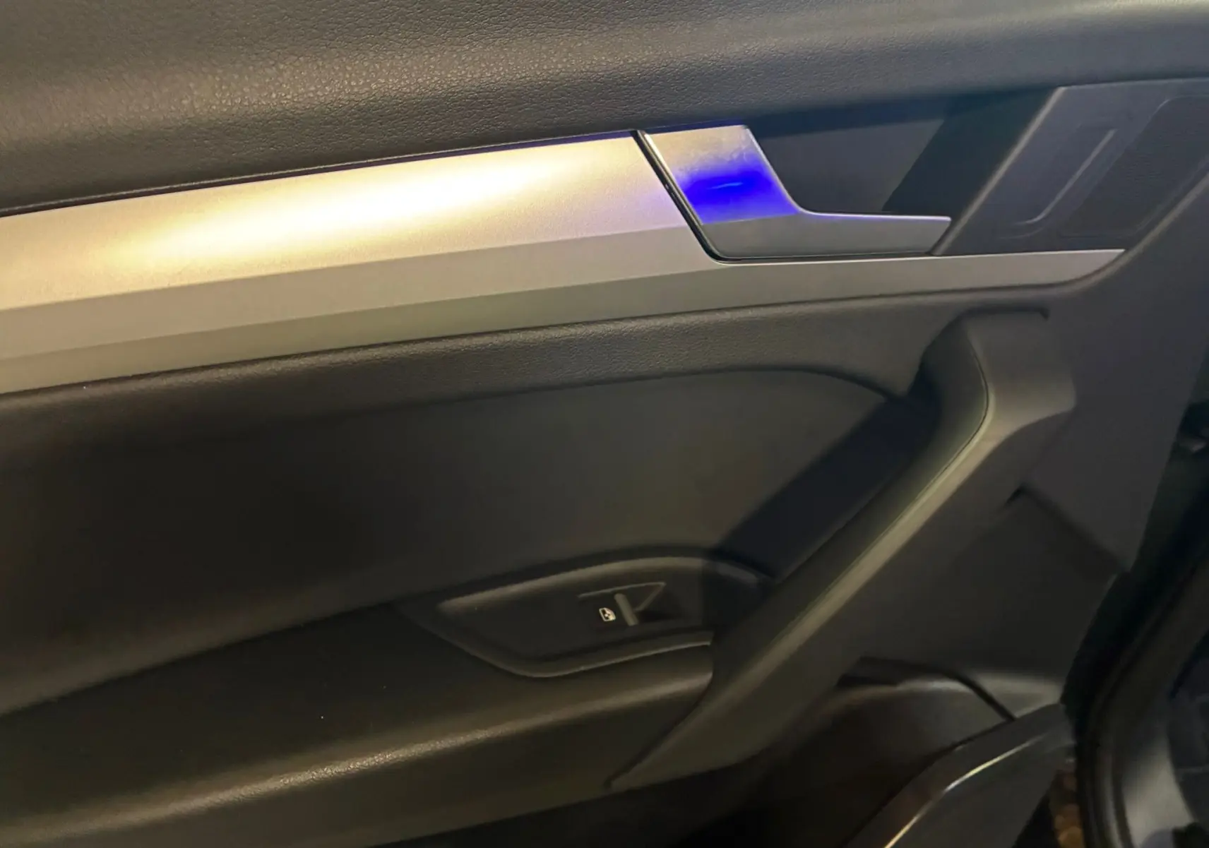 Détail de la porte intérieure côté conducteur de l'Audi Q5 gris 2024 avec poignée argentée et éclairage bleu.