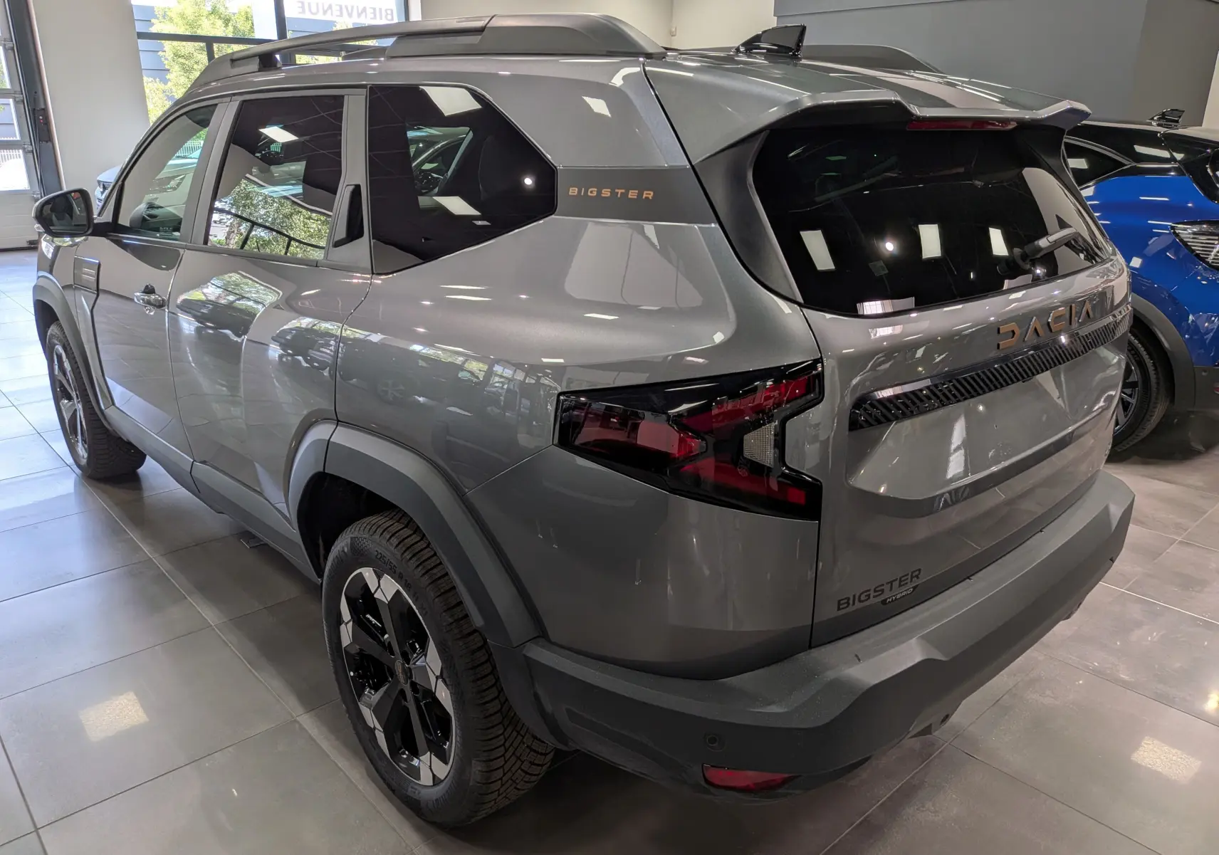Vue 3/4 arrière droite du Dacia Bigster Hybrid-G 150 gris schiste avec logo et feux arrière distinctifs en intérieur showroom.