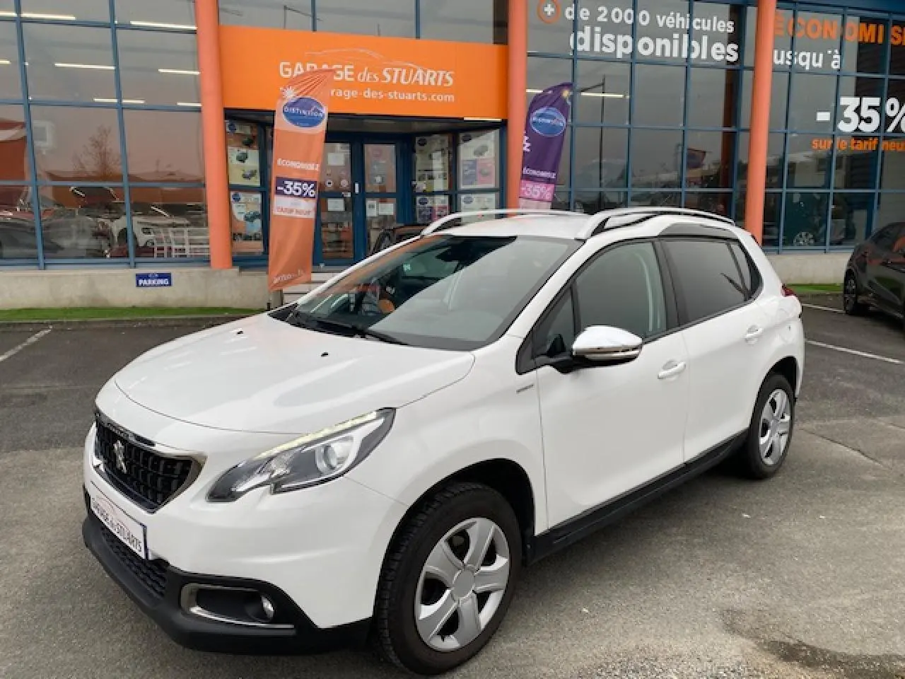 Peugeot 2008 blanc vue 3/4 avant droit, avec jantes acier et barres de toit, stationné devant un garage orange.