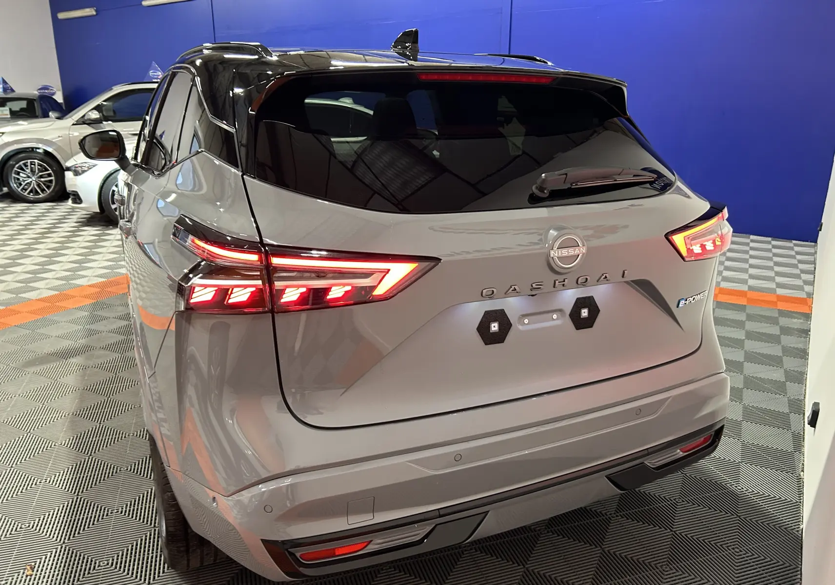 Vue 3/4 arrière d'un Nissan Qashqai gris céramique avec toit noir et feux arrière LED allumés dans un showroom.