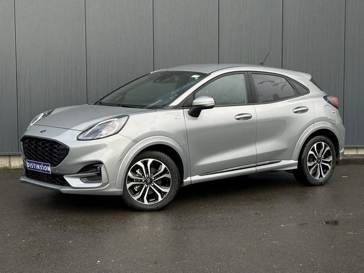 Vue 3/4 avant droit d'un Ford Puma gris Solar avec jantes alliage et vitres surteintées, garé devant un mur gris.