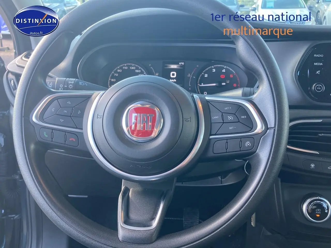 Vue rapprochée du volant multifonction noir avec logo FIAT rouge et tableau de bord numérique d'une Fiat Tipo 2024.
