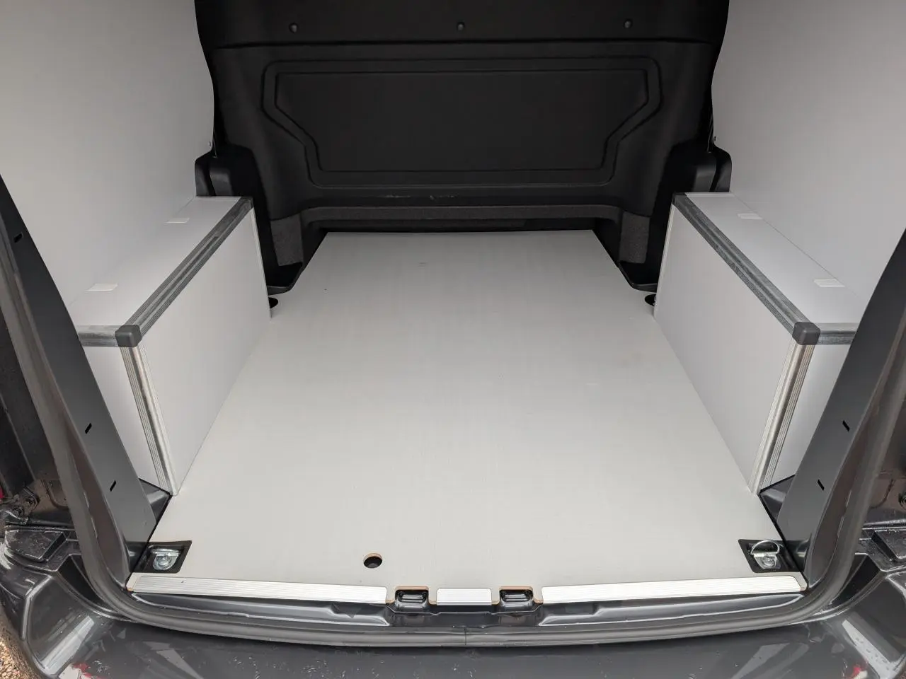 Vue arrière du coffre ouvert du Citroën Jumpy gris, plancher bois clair avec coffrages latéraux blancs.