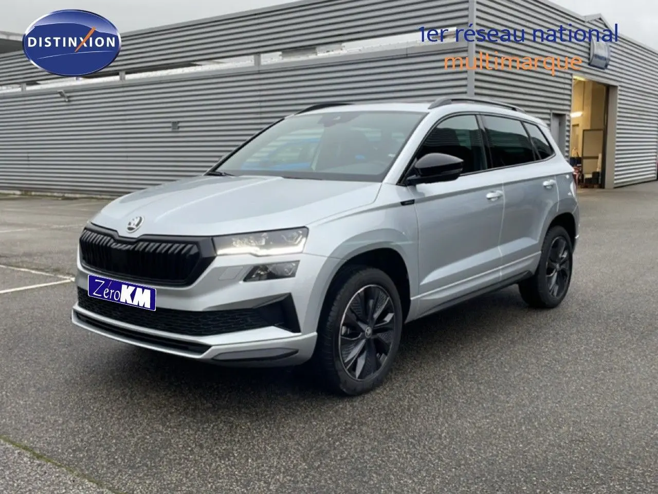 Vue 3/4 avant d'un Skoda Karoq 2025 gris argent métal avec calandre noire et jantes foncées, stationné devant un bâtiment industriel.