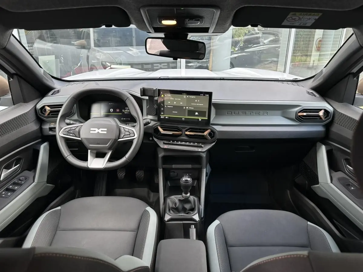 Vue intérieure avant du Dacia Duster blanc glacier 2025, tableau de bord moderne avec écran tactile central et volant multifonction.