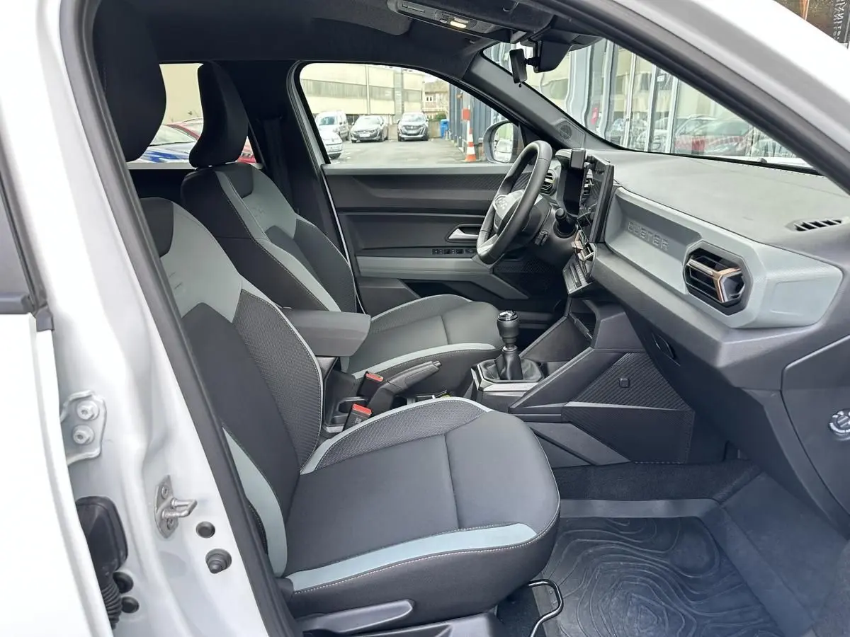 Vue intérieure côté conducteur du Dacia Duster blanc glacier 2025, montrant sièges tissu bicolore et tableau de bord moderne.