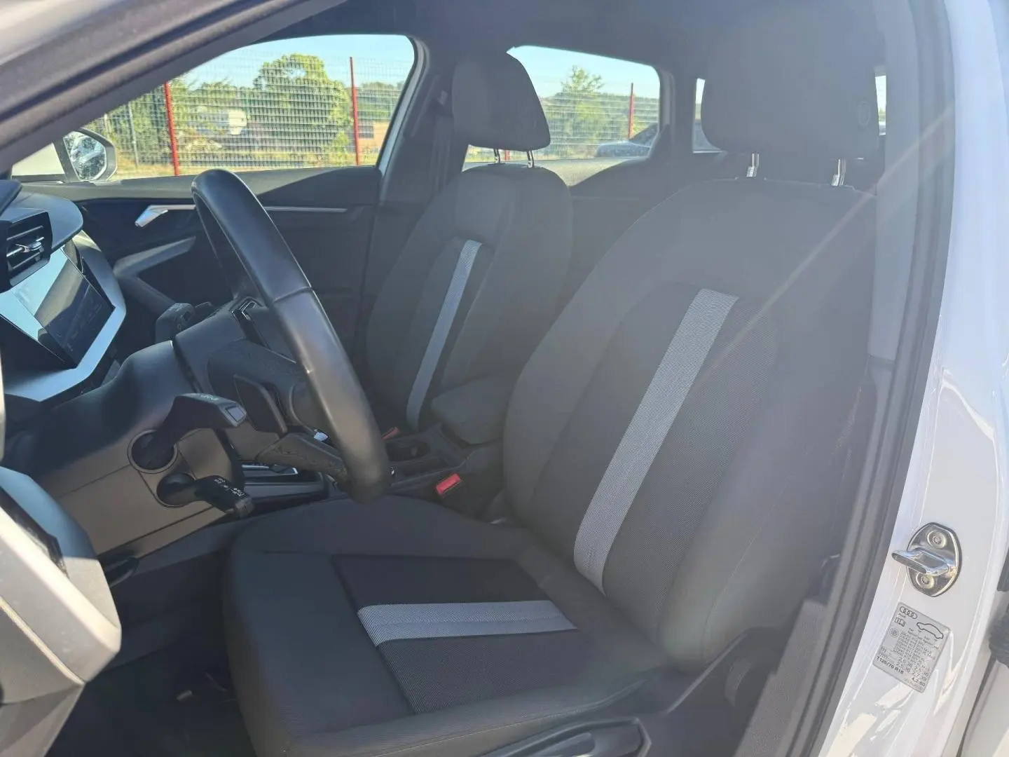 Vue intérieure côté conducteur de l'Audi A3 Sportback blanc Arkona, montrant les sièges tissu noir avec bande centrale grise.
