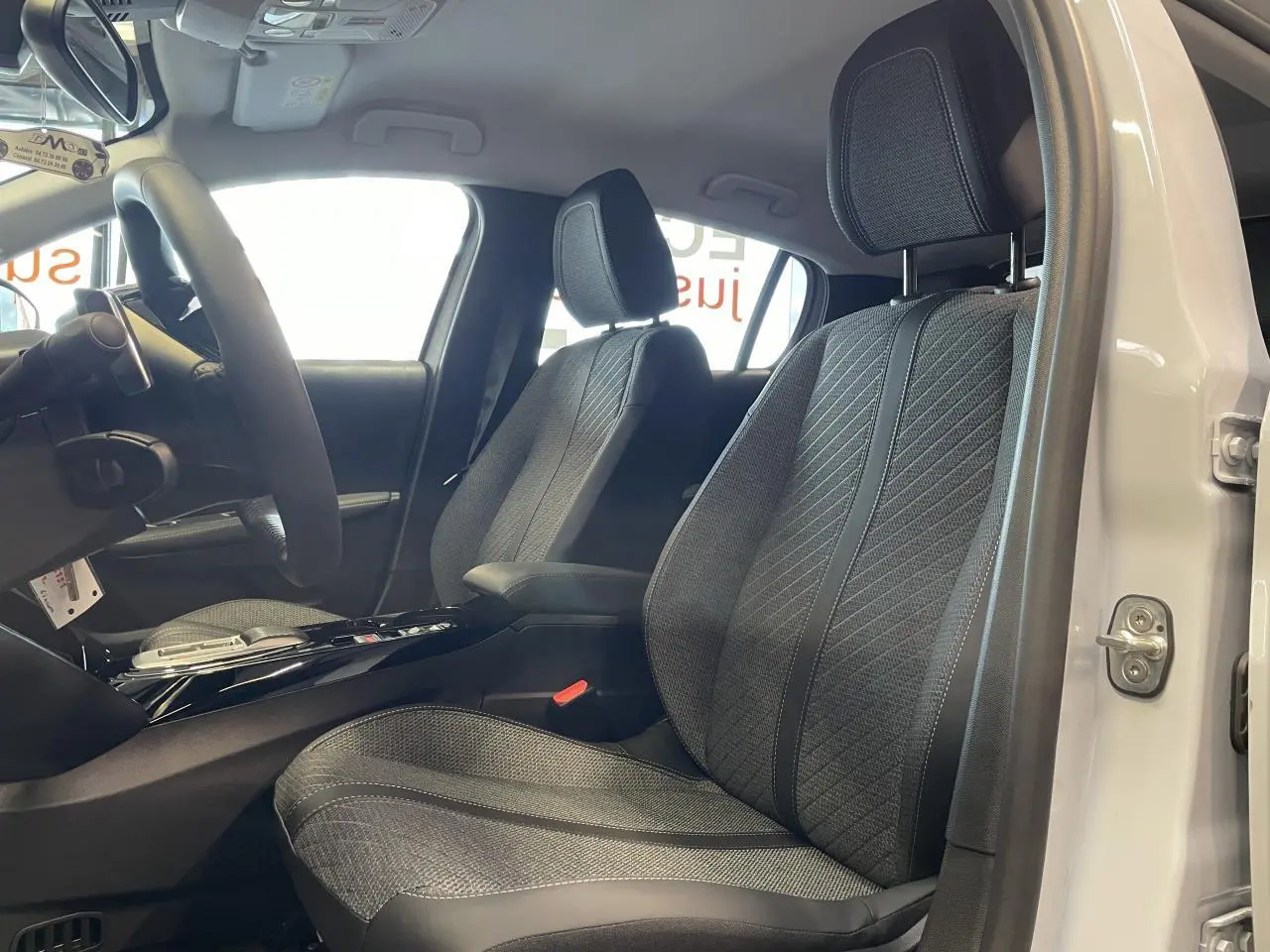Vue intérieure côté conducteur de la PEUGEOT 208 blanche, mettant en valeur les sièges tissu gris et l'accoudoir central.