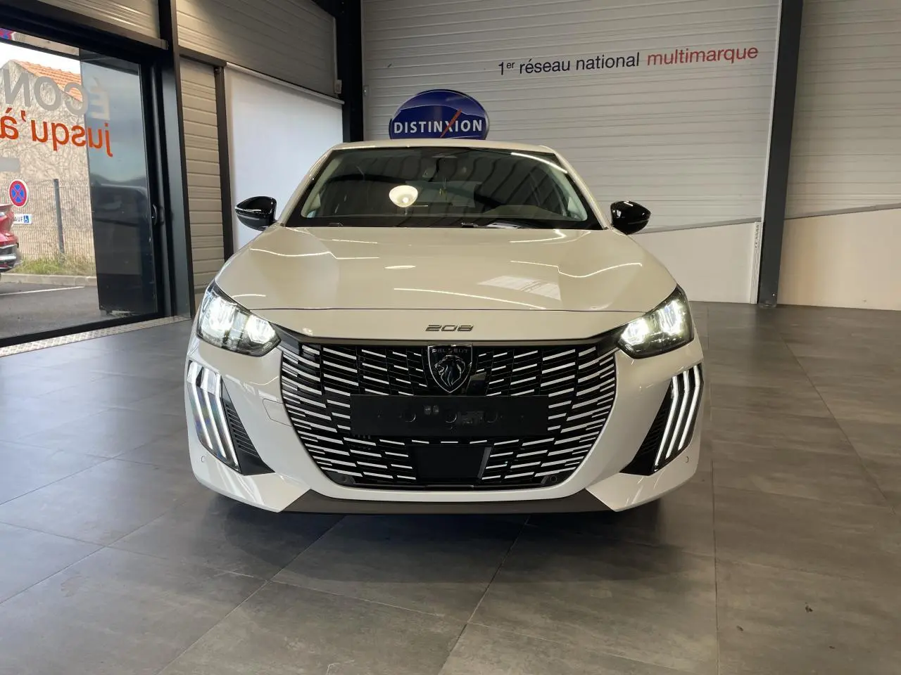 Vue frontale d'une Peugeot 208 blanche Okenite avec calandre noire et feux diurnes à LED en griffes allumés.