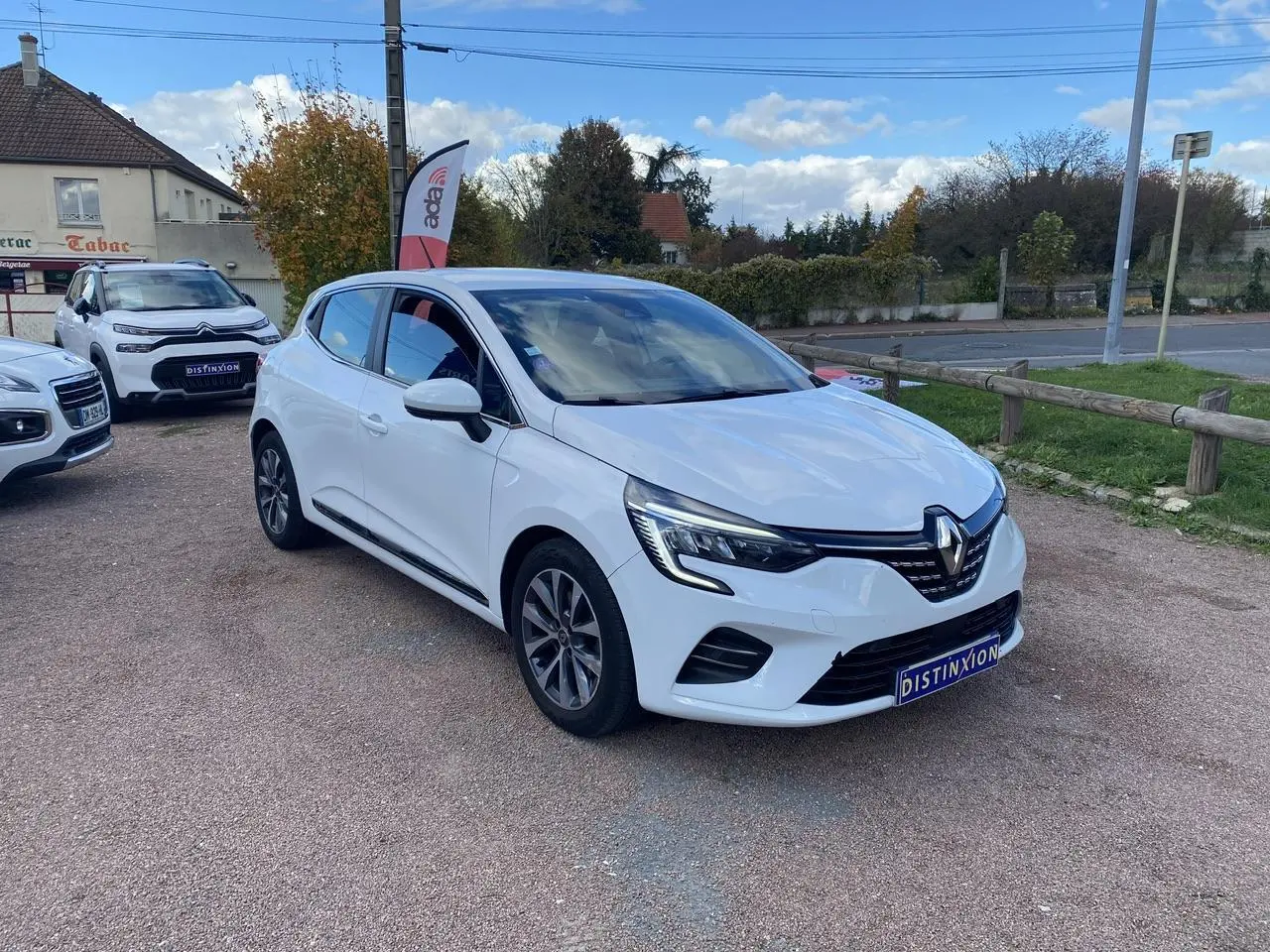 Renault Clio blanc vue 3/4 avant droit, avec phares LED et jantes alliage, stationnée sur un parking extérieur.