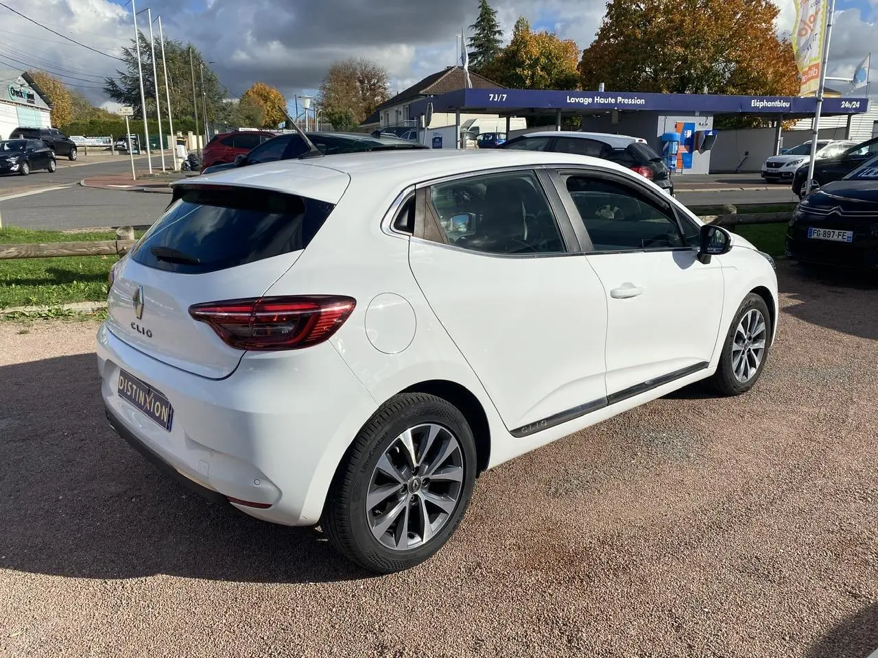 Renault Clio blanche vue 3/4 arrière droit, avec jantes alliage et vitres teintées sur parking extérieur.